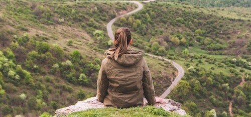 A woman sits on the edge of a cliff looking down at a sloping field of greenery and a road.